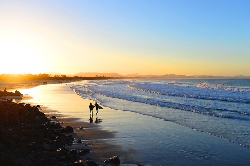 Surfers walk down the beach at sunset in Byron Bay, Australia