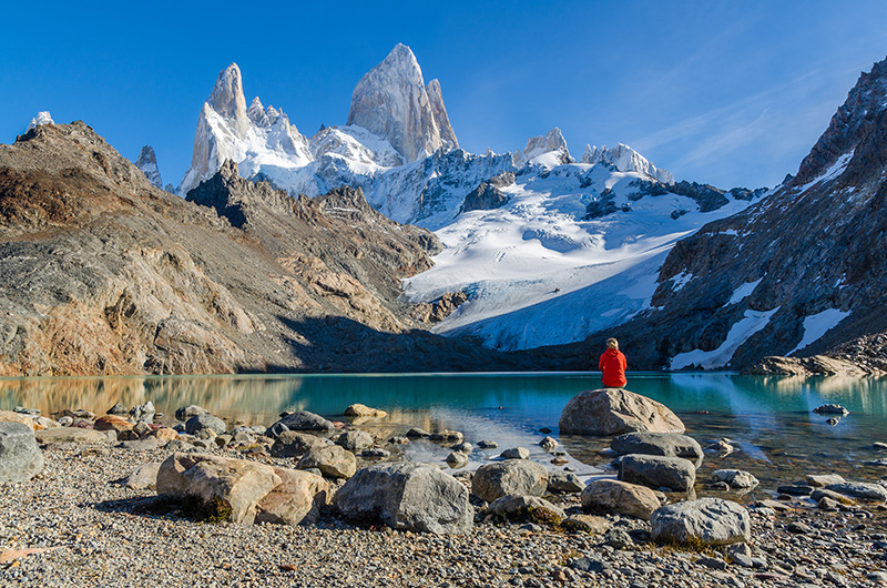 Eco-traveller sits in front of snow-capped mountain and blue lake - Patagonia, Chile